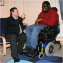 Man sitting in chair speaking with another man sitting in an electric wheelchair
