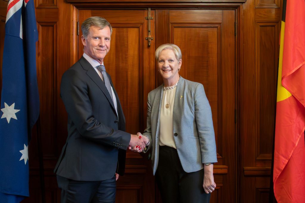 Dan Stubbs shakes hands with The Hon. Maree Edwards MP, Speaker of the Legislative Assembly of Victoria. They are standing in front of a wooden door, flanked by the Australian an Aboriginal flags