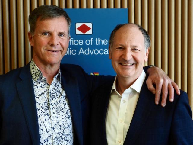 Photograph of Dan Stubbs and Daniel Leighton with their arms around each other standing in front of the Office of the Public Advocate sign